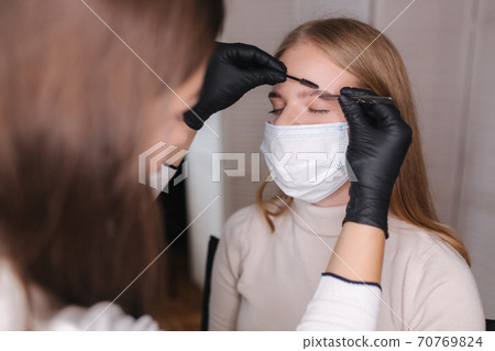 Brow and makeup master in protective mask gives shape to pull out with forceps previously painted with henna eyebrows in a beauty salon. Beautiful blond hair model in medical mask Brow and makeup master in protective mask gives shape to pull out with forceps previously painted with henna eyebrows in a beauty salon. Beautiful blond hair model in medical mask 70769824