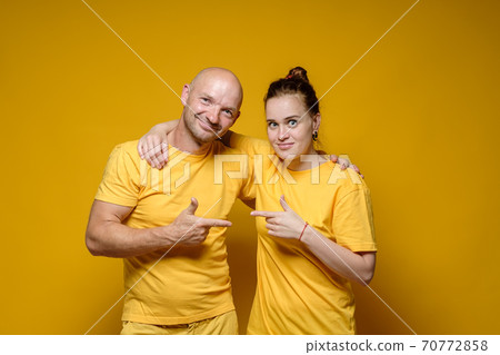 Smiling man and woman in plain, bright clothes hug and point index finger at each other, on a yellow 70772858