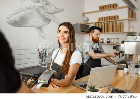 Coffee Business Concept - Beautiful female barista giving payment service for customer with credit card and smiling while working at the bar counter in modern coffee shop. Coffee Business Concept - Beautiful female barista giving payment service for customer with credit card and smiling while working at the bar counter in modern coffee shop. 70775950