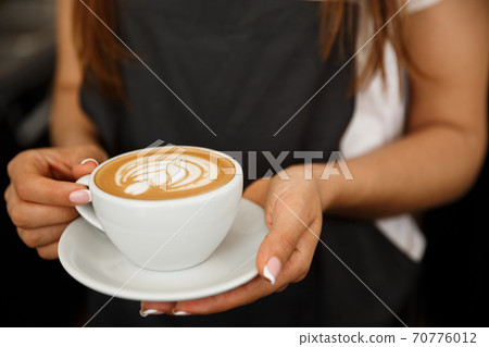 Coffee Business Concept - Cropped Close up of female serving coffee with latte art while standing in coffee shop. Focus on female hands placing a cup of coffee. 70776012