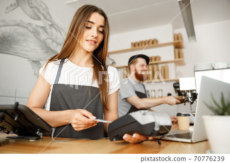 Coffee Business Concept - Beautiful female barista giving payment service for customer with credit card and smiling while working at the bar counter in modern coffee shop. Coffee Business Concept - Beautiful female barista giving payment service for customer with credit card and smiling while working at the bar counter in modern coffee shop. 70776239