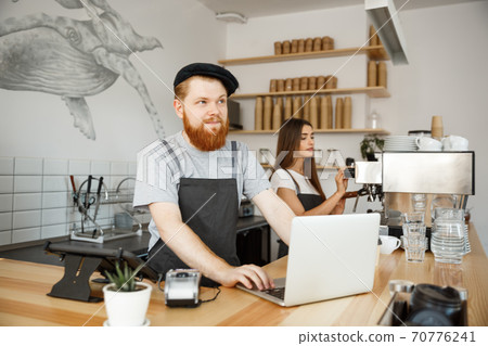 Coffee Business Concept - Young handsome bearded bartende, barista or manager working and planing in laptop at modern coffee shop. Coffee Business Concept - Young handsome bearded bartende, barista or manager working and planing in laptop at modern coffee shop. 70776241