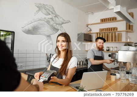 Coffee Business Concept - Beautiful female barista giving payment service for customer with credit card and smiling while working at the bar counter in modern coffee shop. Coffee Business Concept - Beautiful female barista giving payment service for customer with credit card and smiling while working at the bar counter in modern coffee shop. 70776269