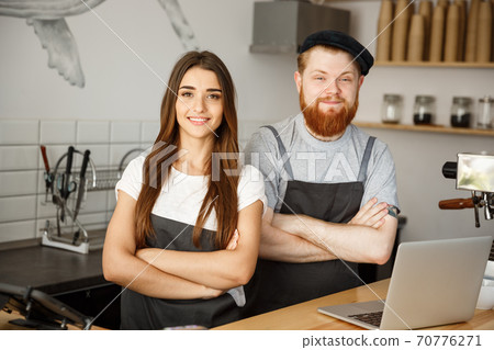 Coffee Business Concept - Positive young bearded man and beautiful attractive lady barista couple in apron looking at camera while standing at bar Couter ready to give Coffee Service at the modern Coffee Business Concept - Positive young bearded man and beautiful attractive lady barista couple in apron looking at camera while standing at bar Couter ready to give Coffee Service at the modern 70776271