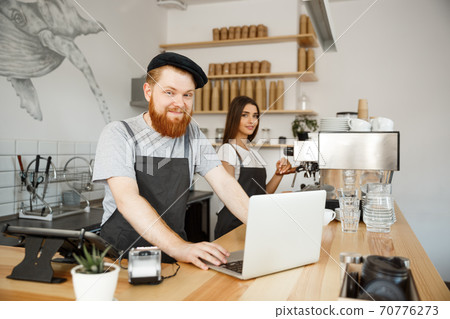 Coffee Business Concept - Young handsome bearded bartender, barista or manager working and planing in laptop at modern coffee shop. Coffee Business Concept - Young handsome bearded bartender, barista or manager working and planing in laptop at modern coffee shop. 70776273