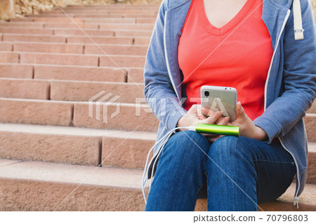A girl sitting on the steps uses a smartphone outdoors while charging from an external power bank A girl sitting on the steps uses a smartphone outdoors while charging from an external power bank 70796803