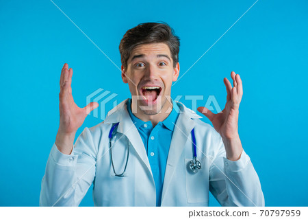 Amazed doctor man shocked, saying WOW. Handsome doc in medical coat surprised to camera over blue 70797959