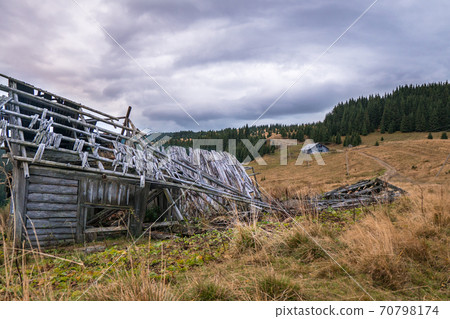 Old destroyed settlement in the mountains among the pastures with dried grass and fir-trees forest on the background. Old destroyed settlement in the mountains among the pastures with dried grass and fir-trees forest on the background. 70798174