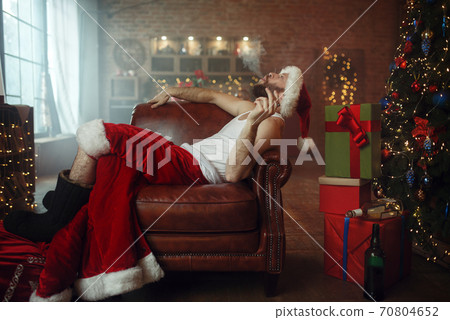 Vile Santa claus in red cap smoking cigar on couch Vile Santa claus in red cap smoking cigar on couch 70804652