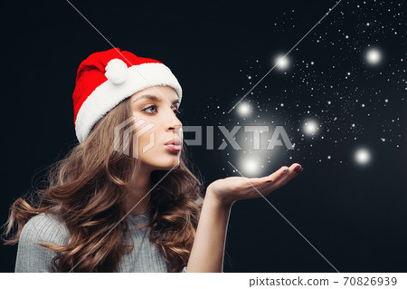 Beautiful girl in Santa hat blowing on hand while Beautiful girl in Santa hat blowing on hand while 70826939
