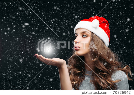 Beautiful girl in Santa hat blowing on hand while Beautiful girl in Santa hat blowing on hand while 70826942