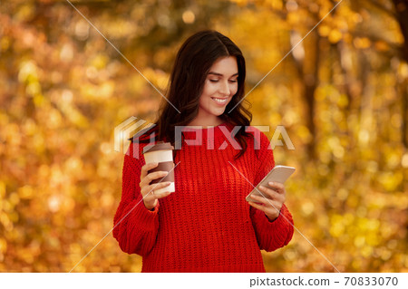 Lovely young lady in in warm knitted sweater using her smartphone and drinking takeout coffee at autumn park Lovely young lady in in warm knitted sweater using her smartphone and drinking takeout coffee at autumn park 70833070