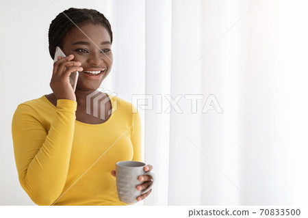 Positive African Lady Talking On Cellphone And Enjoying Morning Coffee Near Window Positive African Lady Talking On Cellphone And Enjoying Morning Coffee Near Window 70833500