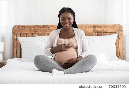 Home Portrait Of Beautiful Pregnant African Woman Sitting On Bed Embracing Belly 70833503