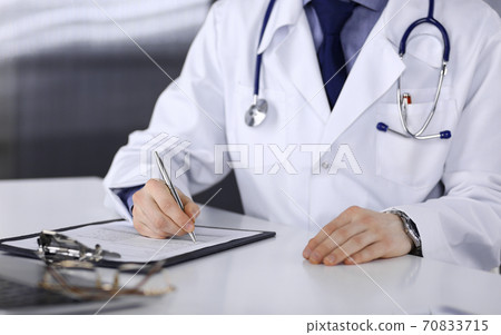 Unknown male doctor sitting and working with clipboard of medication history record in clinic at his working place, close-up. Young physician at work. Perfect medical service, medicine concept 70833715