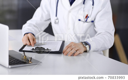 Unknown male doctor sitting and working with clipboard of medication history record in clinic at his working place, close-up. Young physician at work. Perfect medical service, medicine concept Unknown male doctor sitting and working with clipboard of medication history record in clinic at his working place, close-up. Young physician at work. Perfect medical service, medicine concept 70833993