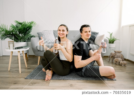 Family sport and healthy lifestyle concept. Young couple in lotus pose, sitting on the floor in front of stylish gray sofa, resting after workout, holding bottles of water and showing thumbs up 70835019