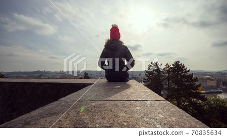 Woman in winter jacket sitting on a wall and looking at the panorama of Prague Woman in winter jacket sitting on a wall and looking at the panorama of Prague 70835634