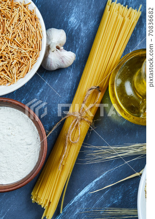 Various uncooked pasta with a bottle of oil, flour, and garlic on a blue background Various uncooked pasta with a bottle of oil, flour, and garlic on a blue background 70836682