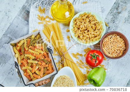 Bowls of uncooked pasta, spaghetti, a basket of fusilli, vegetables, and a bottle of oil on a marble background 70836718