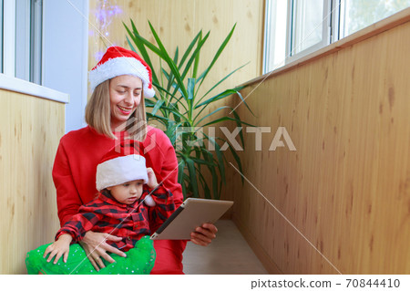 christmas online congratulations: a woman with a child in red Santa hats congratulating family or friends with a tablet. the mother and daughter celebrates the new year in isolation. Winter holidays christmas online congratulations: a woman with a child in red Santa hats congratulating family or friends with a tablet. the mother and daughter celebrates the new year in isolation. Winter holidays 70844410