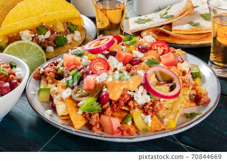 Mexican food. Nachos with beef, beans, chili peppers, onions and cheese sauce close-up, with pork tacos, quesadillas and tequila in the background 70844669
