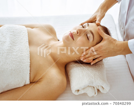 Beautiful brunette woman enjoying facial massage with closed eyes. Relaxing treatment in medicine and spa center concepts Beautiful brunette woman enjoying facial massage with closed eyes. Relaxing treatment in medicine and spa center concepts 70848299