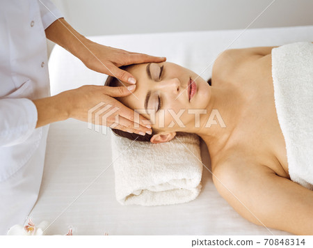 Beautiful brunette woman enjoying facial massage with closed eyes. Relaxing treatment in medicine and spa center concepts 70848314