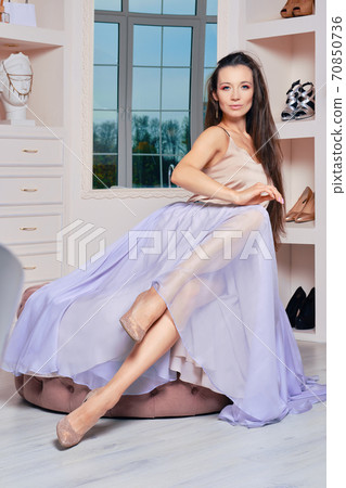 Woman in long satin skirt and blouse sit on ottoman at home Woman in long satin skirt and blouse sit on ottoman at home 70850736