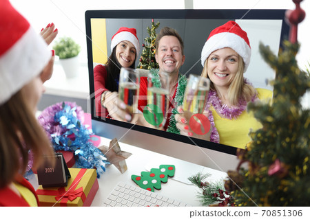 Two women and man in santa claus hats with glasses of champagne in hands congratulate friend happy new year and merry christmas through computer screen 70851306