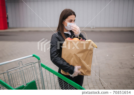 Young woman in protective mask and disposable gloves carries a craft package with groceries to her car in front of supermarket 70851654