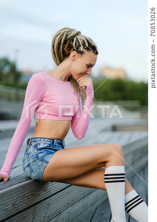 Summer urban portrait of a slender young beautiful woman with dreadlocks in a pink T-shirt and denim short shorts on a wooden staircase. 70853026