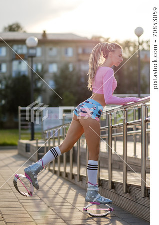 Young beautiful sportswoman with dreadlocks, in bright sportswear. The girl is engaged in sports on the city sports ground. Kangoo jumps. 70853059
