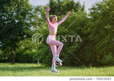 A young slender beautiful woman with dreadlocks in sportswear trains in the park outdoors. Kangoo jumps training. 70853085