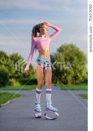 A young slender beautiful woman with dreadlocks in sportswear trains in the park outdoors. Kangoo jumps training. 70853090
