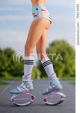 A young slender beautiful woman with dreadlocks in sportswear trains in the park outdoors. Kangoo jumps training. 70853095
