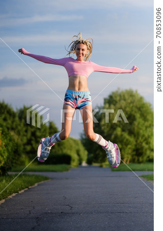A young slender beautiful woman with dreadlocks in sportswear trains in the park outdoors. Kangoo jumps training. 70853096