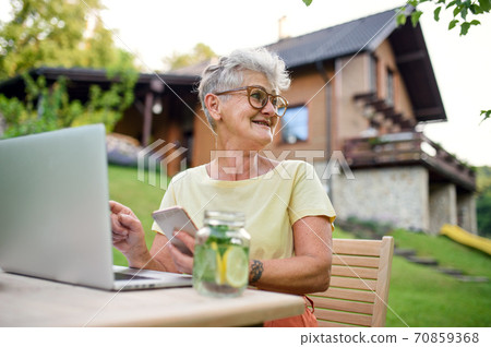 Senior woman with laptop and smartphone working outdoors in garden, home office concept. 70859368
