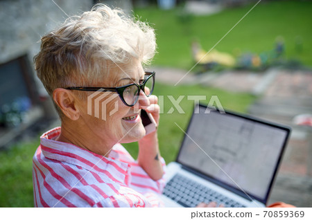 Senior woman architect with smartphone working outdoors in garden, home office concept. 70859369