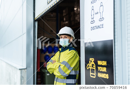 Woman worker with face mask standing in front of warehouse, coronavirus concept. Woman worker with face mask standing in front of warehouse, coronavirus concept. 70859416