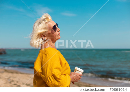 An adult woman in sunglasses and a yellow blouse with a paper cup of coffee to go by the sea on a sunny day. Coffee to go An adult woman in sunglasses and a yellow blouse with a paper cup of coffee to go by the sea on a sunny day. Coffee to go 70861014