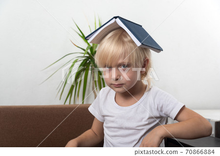 Fair-haired boy sits with book on his head. Angry little boy holding book above his head like roof Fair-haired boy sits with book on his head. Angry little boy holding book above his head like roof 70866884