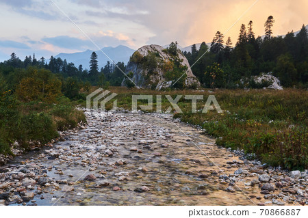 evening wooded mountain valley with blurred river water and a scenic cliff in the distance evening wooded mountain valley with blurred river water and a scenic cliff in the distance 70866887