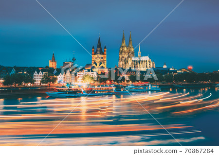 Cologne, Germany. Great St. Martin Church And Dom In Cologne At Evening With Reflection In River Rhine. Evening Blue Hour. Light Trails On Water From Pleasure Boats In Long Exposure Photography. Cologne, Germany. Great St. Martin Church And Dom In Cologne At Evening With Reflection In River Rhine. Evening Blue Hour. Light Trails On Water From Pleasure Boats In Long Exposure Photography. 70867284