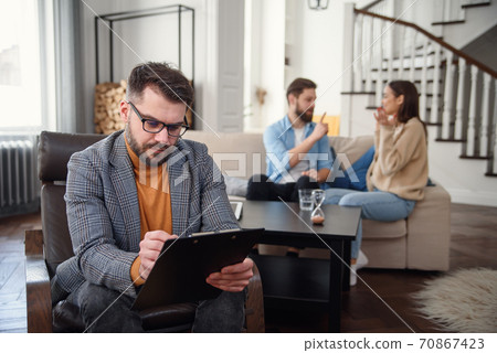 Depressed couple is sitting on couch and quarrelling to each other while psychologist doctor is making notes. Depressed couple is sitting on couch and quarrelling to each other while psychologist doctor is making notes. 70867423