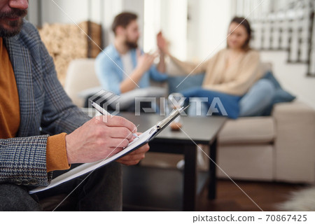 Beautiful young couple is sitting on couch and talking to the psychologist while doctor is making notes Beautiful young couple is sitting on couch and talking to the psychologist while doctor is making notes 70867425
