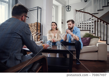 Close up of male psychologist sits at chair and listens depressed young couple at stylish cozy office. Selective focus. 70867497