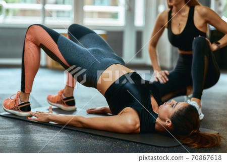 Staying fit. Young caucasian woman in sportswear lying on yoga mat at gym and doing abs exercises with assistance of personal trainer or fitness instructor 70867618