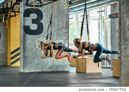 Two young athletic girls exercising with trx fitness straps at gym 70867750