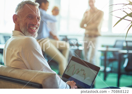 Young cheerful mature businessman sitting in the modern office, using laptop and smiling at camera while his colleagues talking on the background Young cheerful mature businessman sitting in the modern office, using laptop and smiling at camera while his colleagues talking on the background 70868122